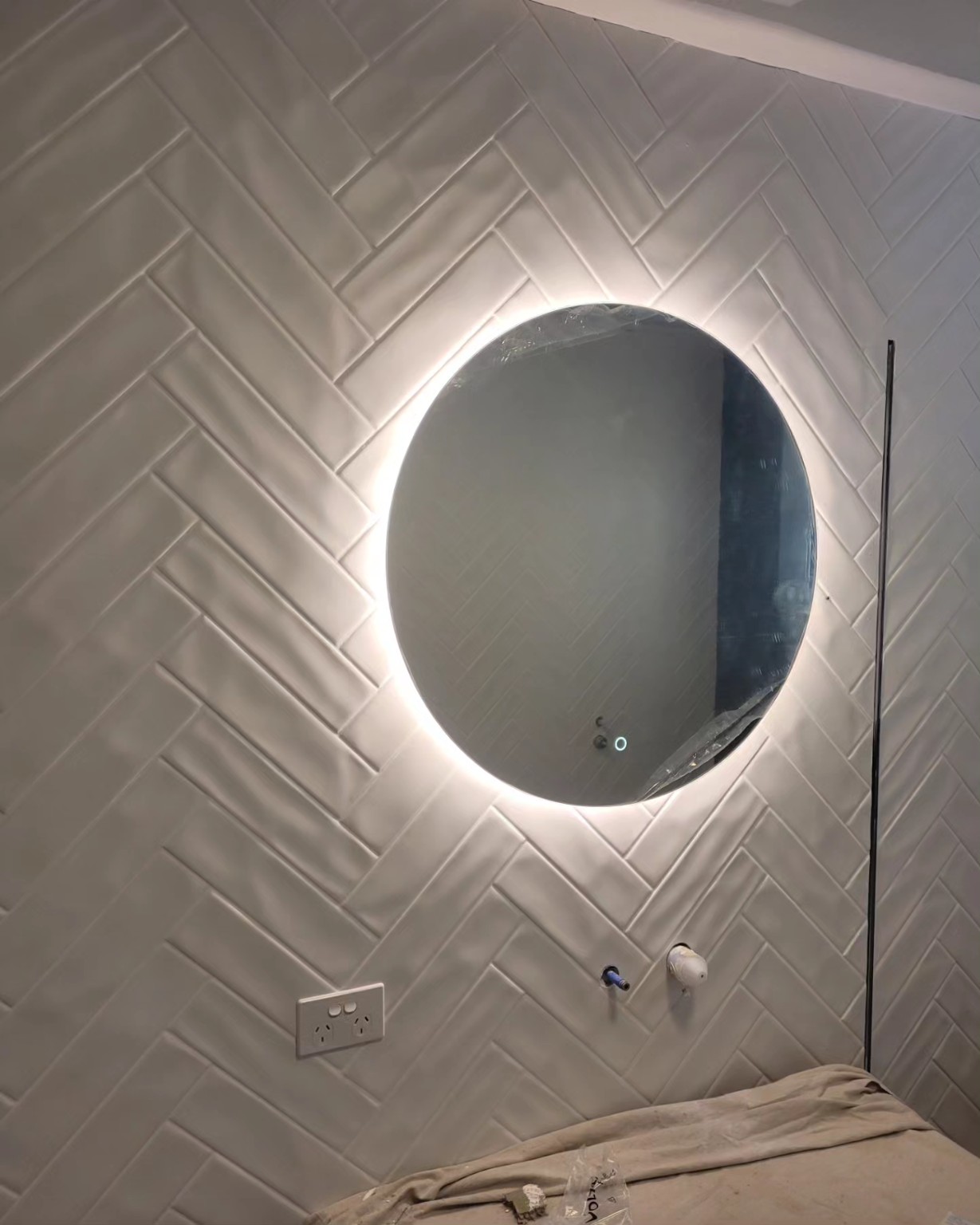 Round illuminated bathroom mirror installed against herringbone tiles